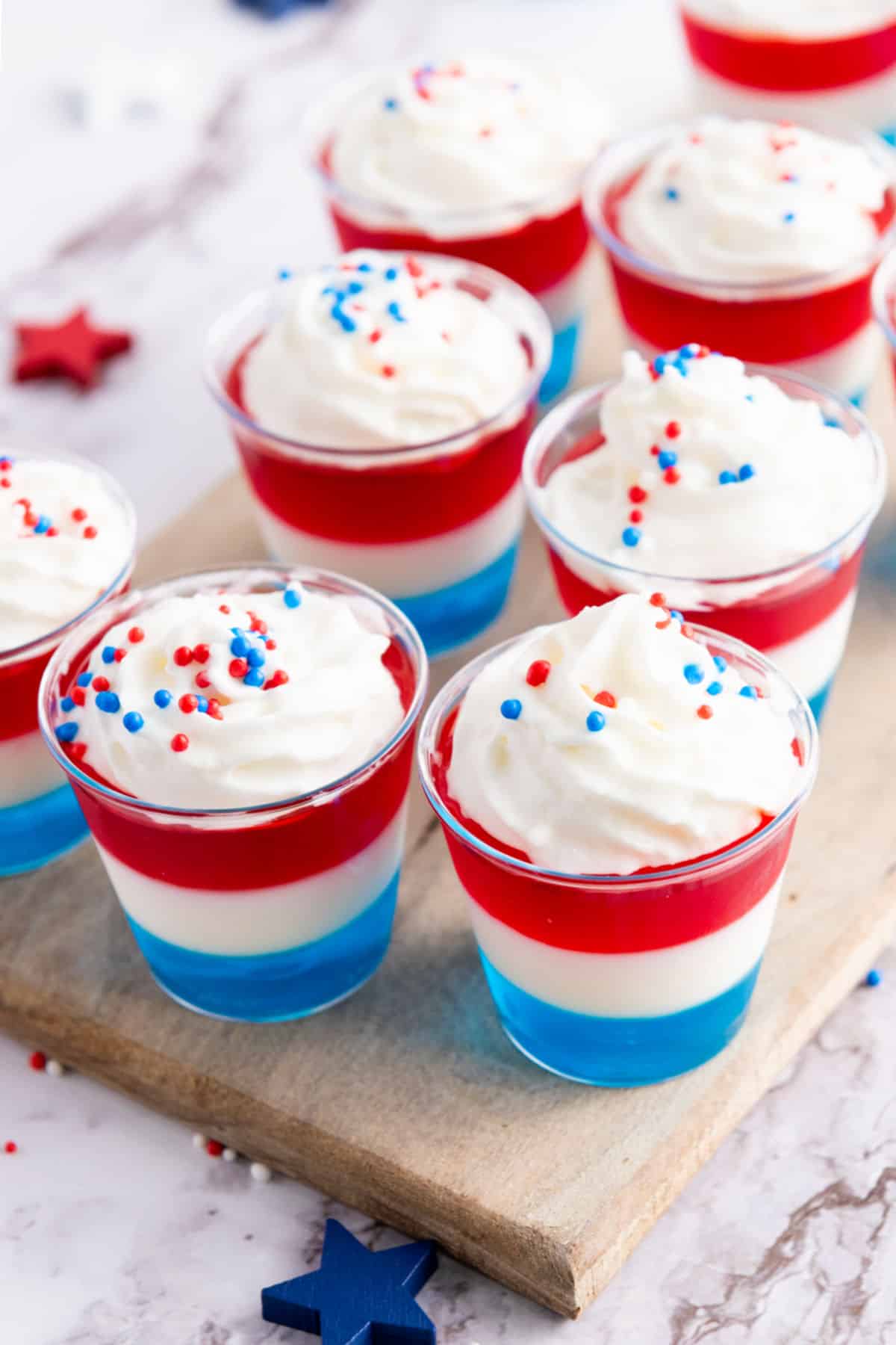 Patriotic cream and sprinkle-topped jello cups on a wooden board.