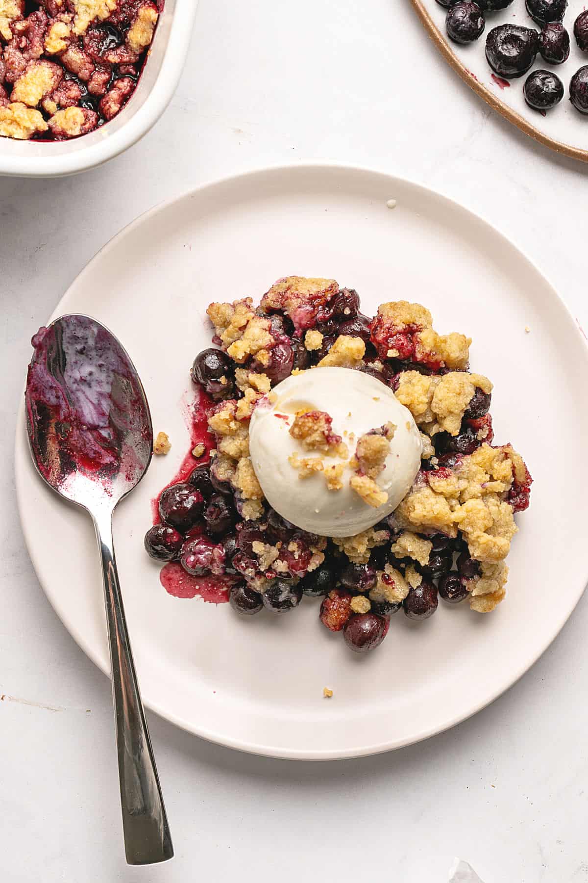 Serving of blueberry crumble with ice cream. White plate and background, silver spoon.