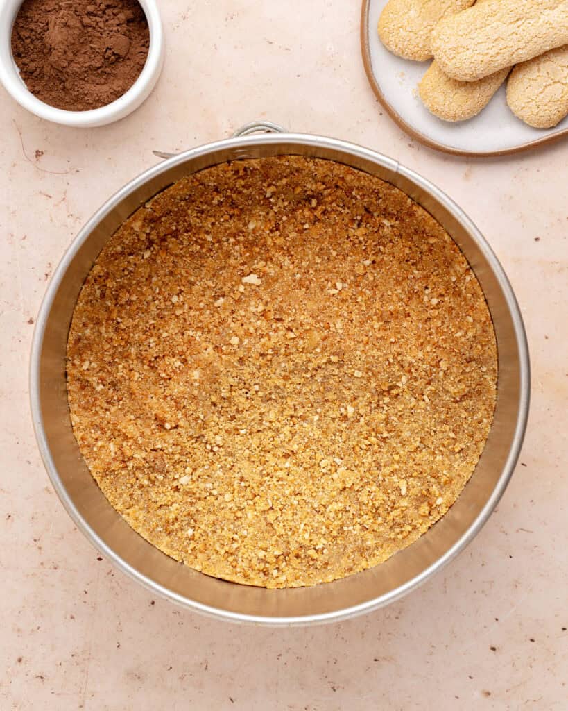 Round metal pan with crumb crust. Light peach surface, plate of ladyfingers and bowl with cocoa powder.