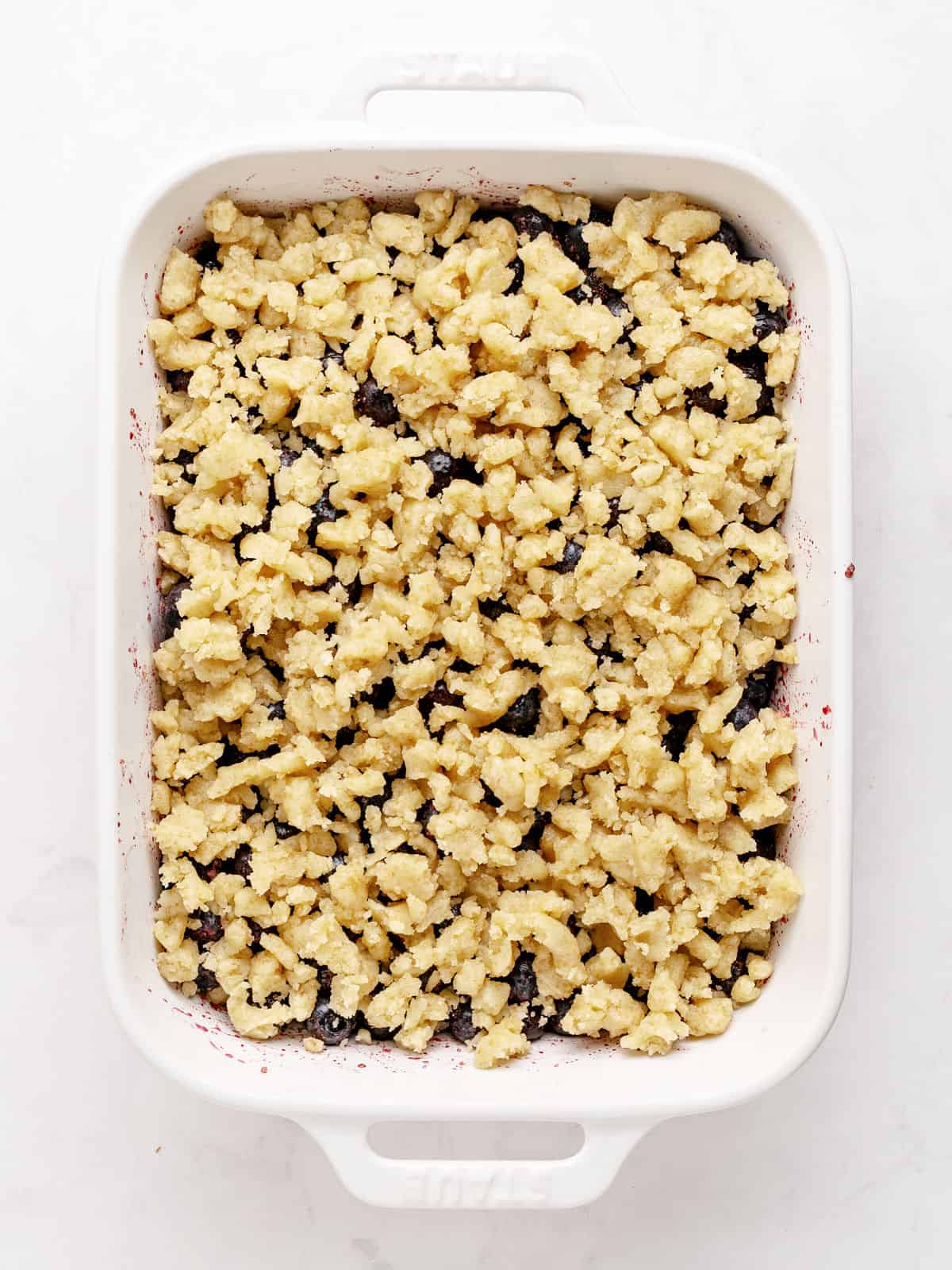 Rectangular white ceramic dish with blueberry crumble before baking. White marble surface.