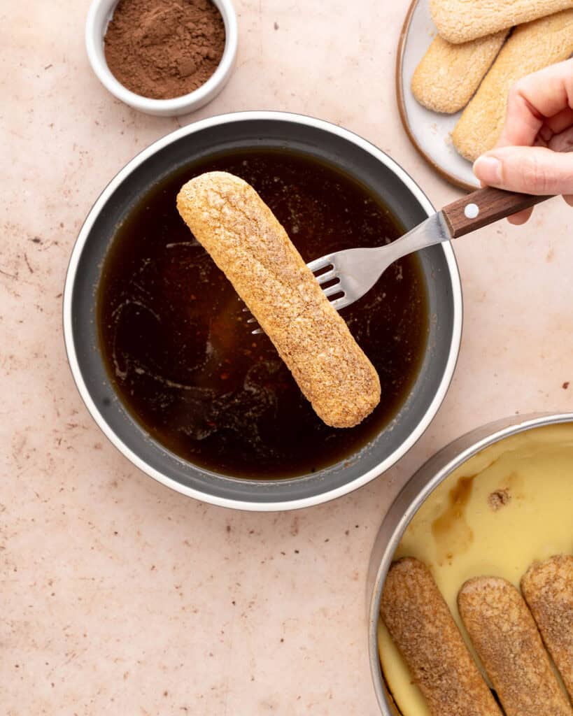 Fork holding ladyfinger over bowl with coffee. Peach surface. Half-assembled tiramisu cheesecake.