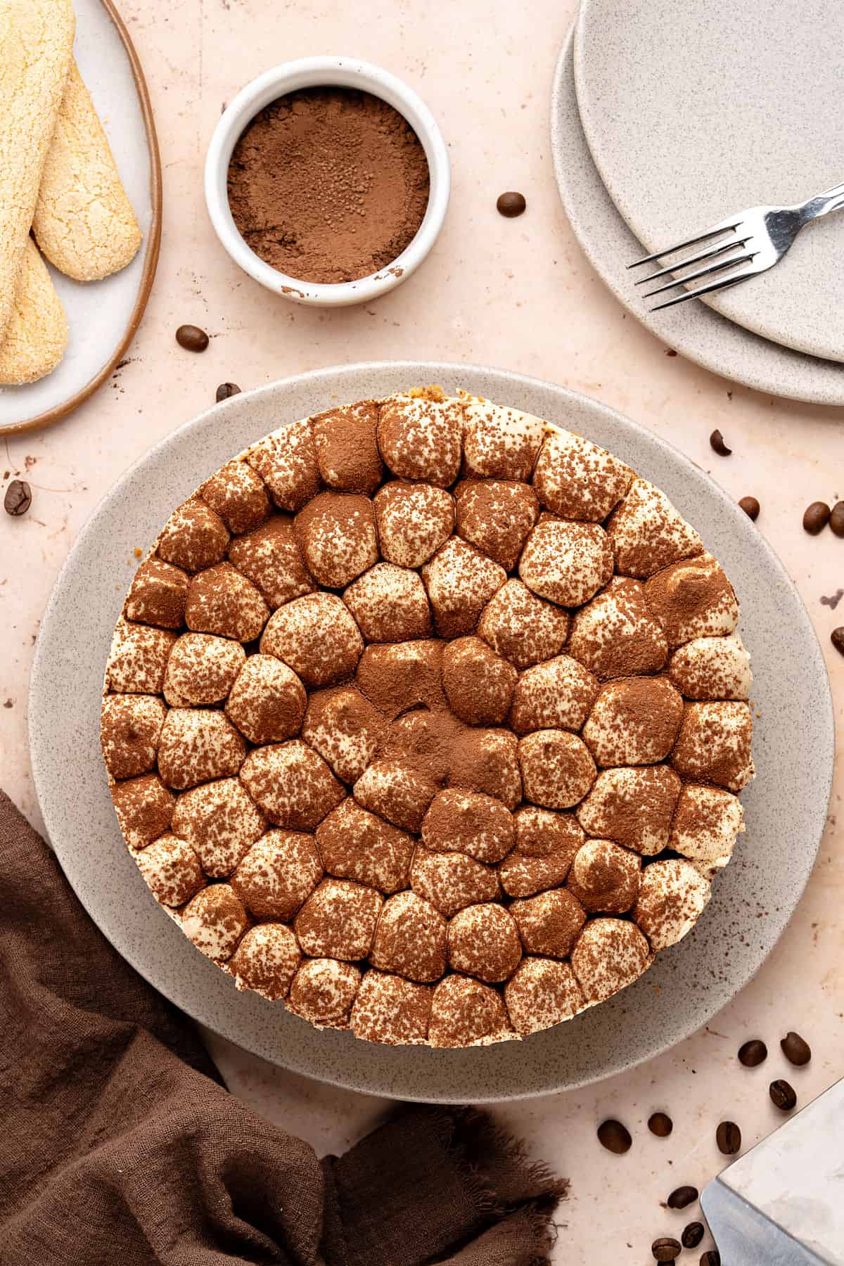 Whole cream-cocoa topped tiramisu cheesecake on a gray plate. Top view. Bowl with coffee, light peach background, coffee beans, brown towel.
