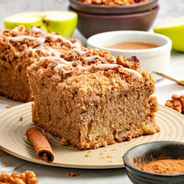 Square of applesauce coffee cake on a beige plate. Cinnamon stick and bowl, apple slices.