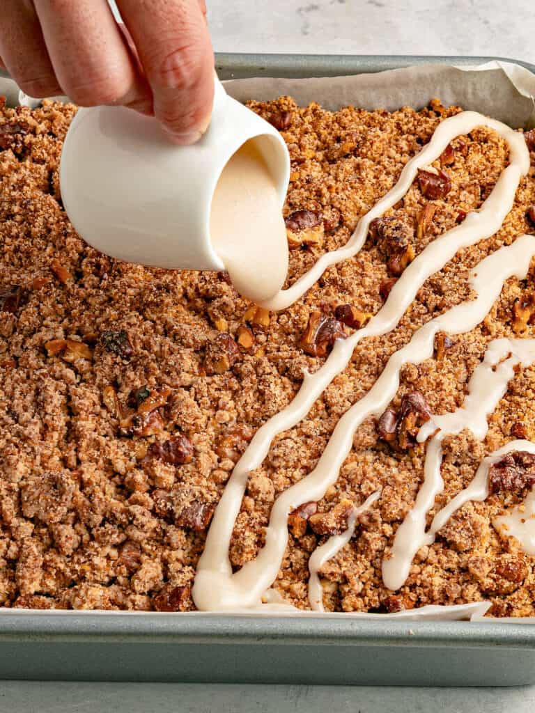 Pouring glaze from a white jar on a crumb cake in a metal pan.