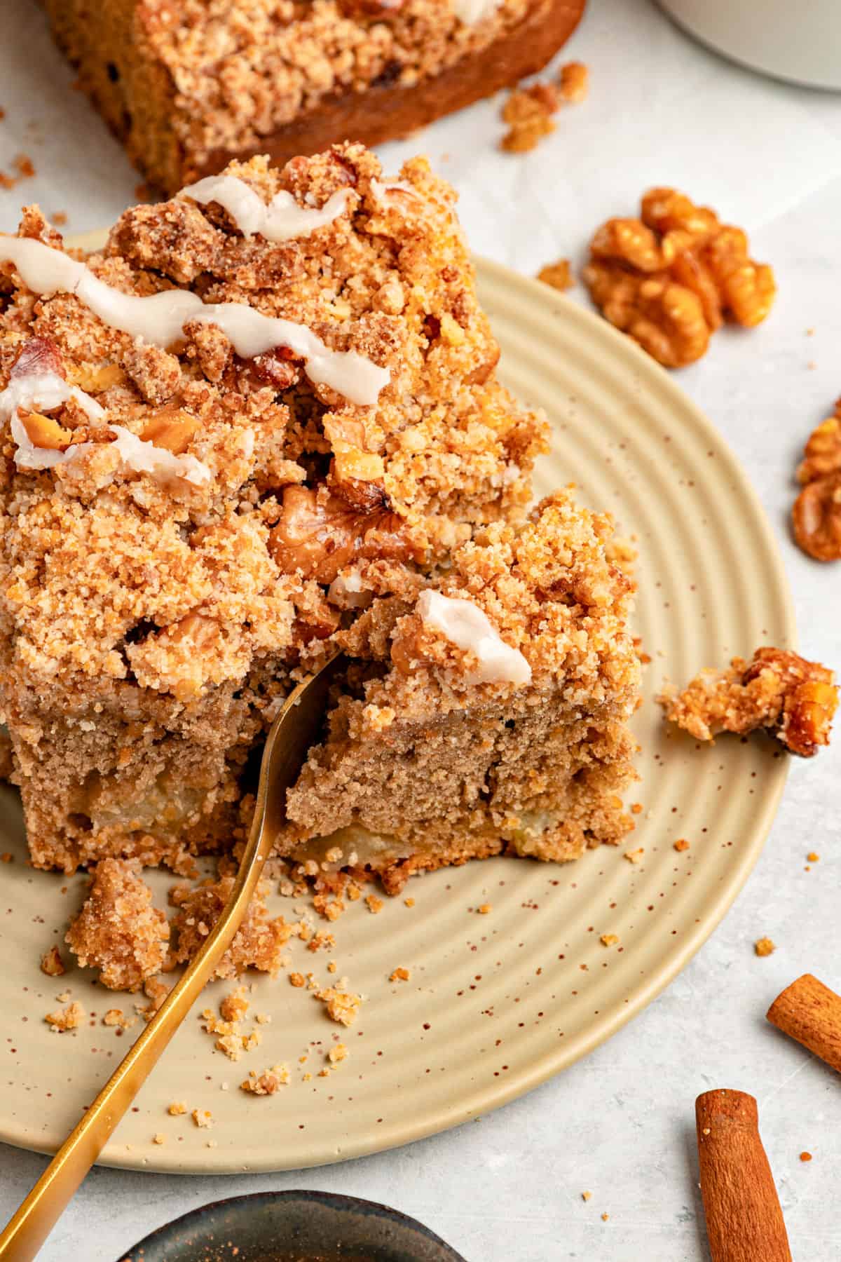 Forking a slice of applesauce crumb cake on a beige plate. Pecans, light gray surface, gold fork.