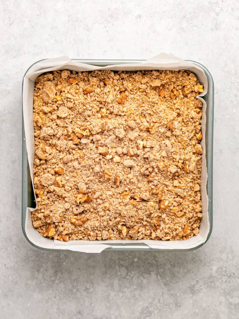 Walnut crumble topped cake in a parchment-lined square metal pan. Light gray surface.
