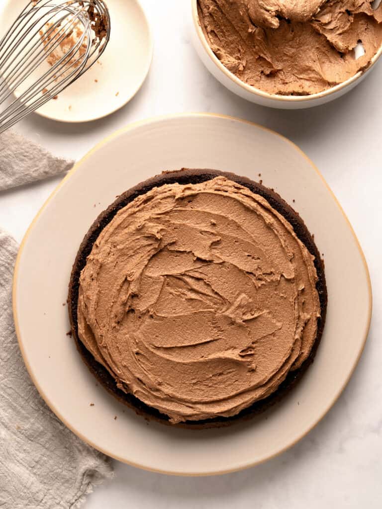 Filling round of chocolate cake with chocolate buttercream. Whitish plate, bowl with frosting, wire whisk.