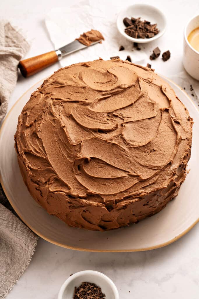 Whole frosted chocolate cake on a whitish plate. Top view. Beige linen, butter knife, white bowls.