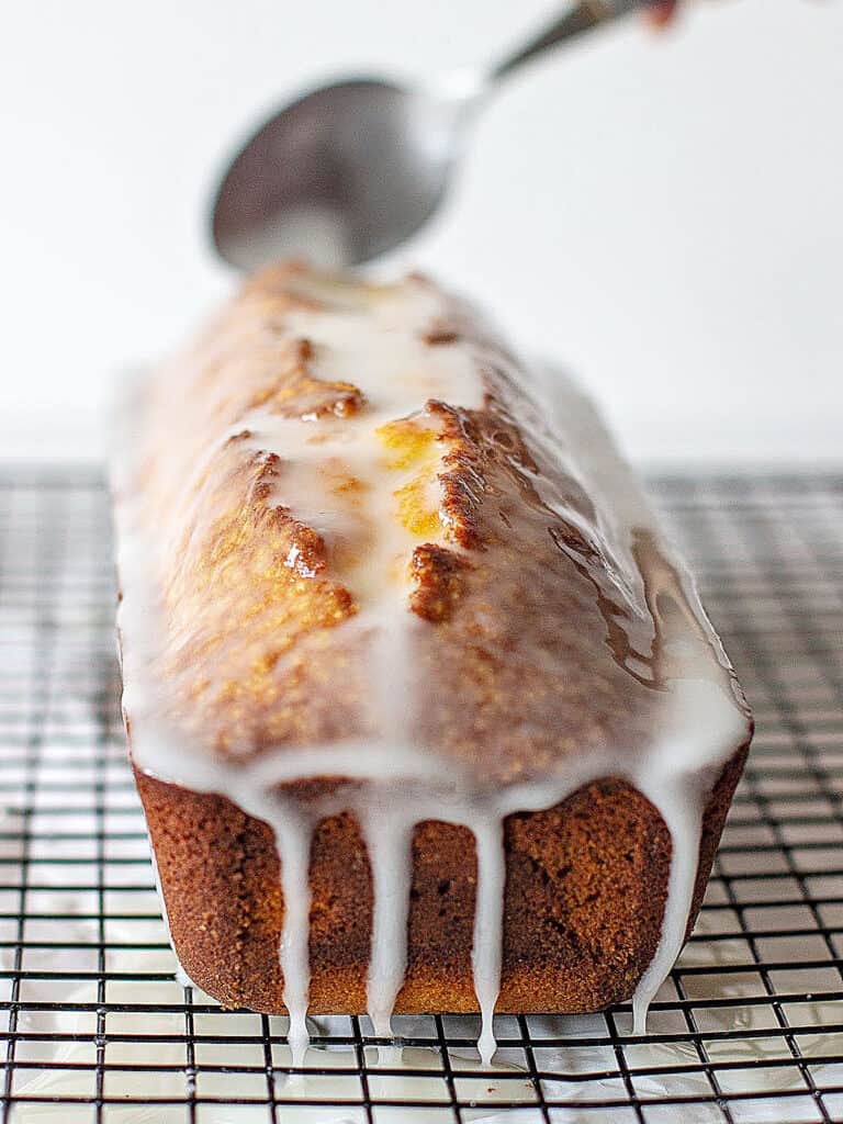 Glazing a loaf cake with a spoon on a black wire rack. White background.