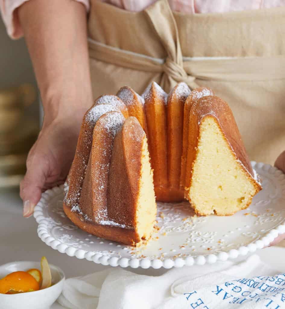 Person in beige apron holding a white plate with a plain bundt cake. Slices missing.