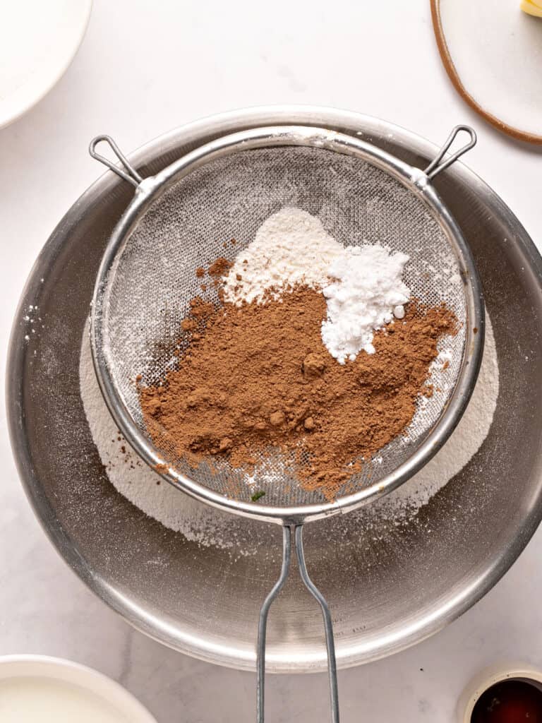Sifting cocoa powder and dry ingredients over a metal bowl. White surface. Top view.