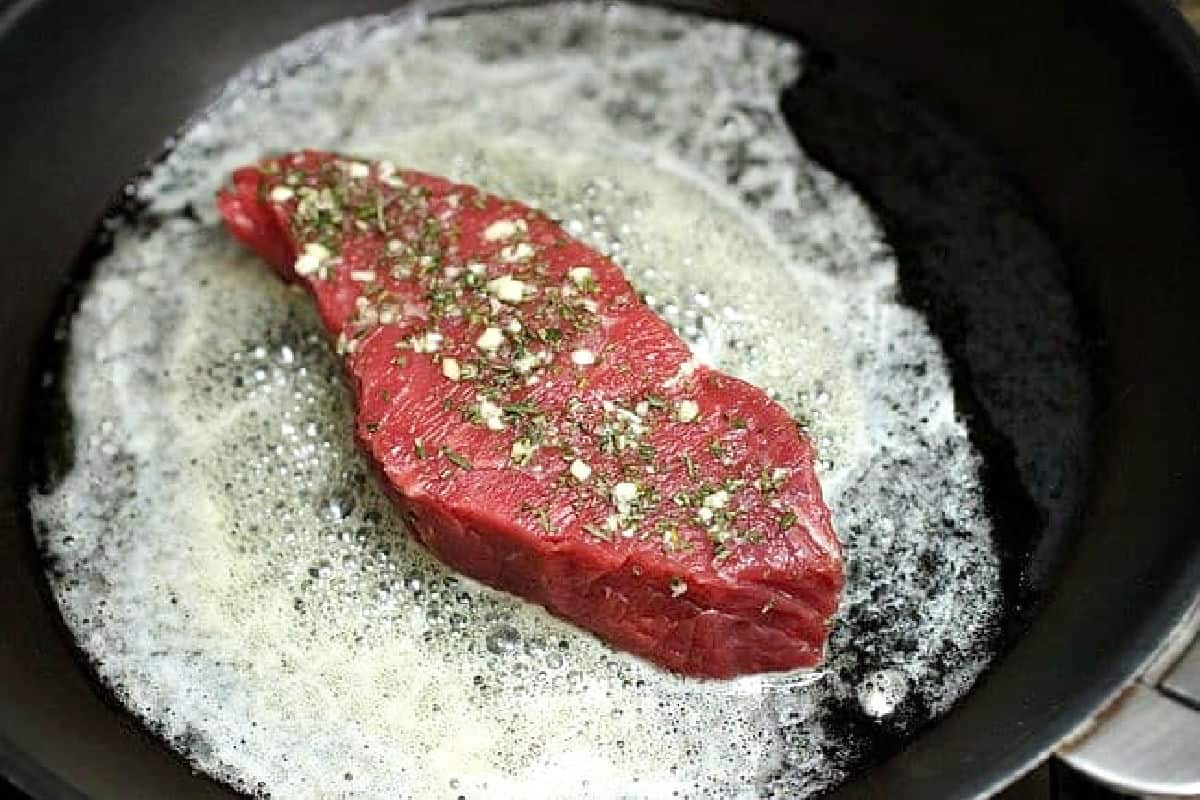 Raw steak with rosemay and garlic cooked in butter on a black pan.