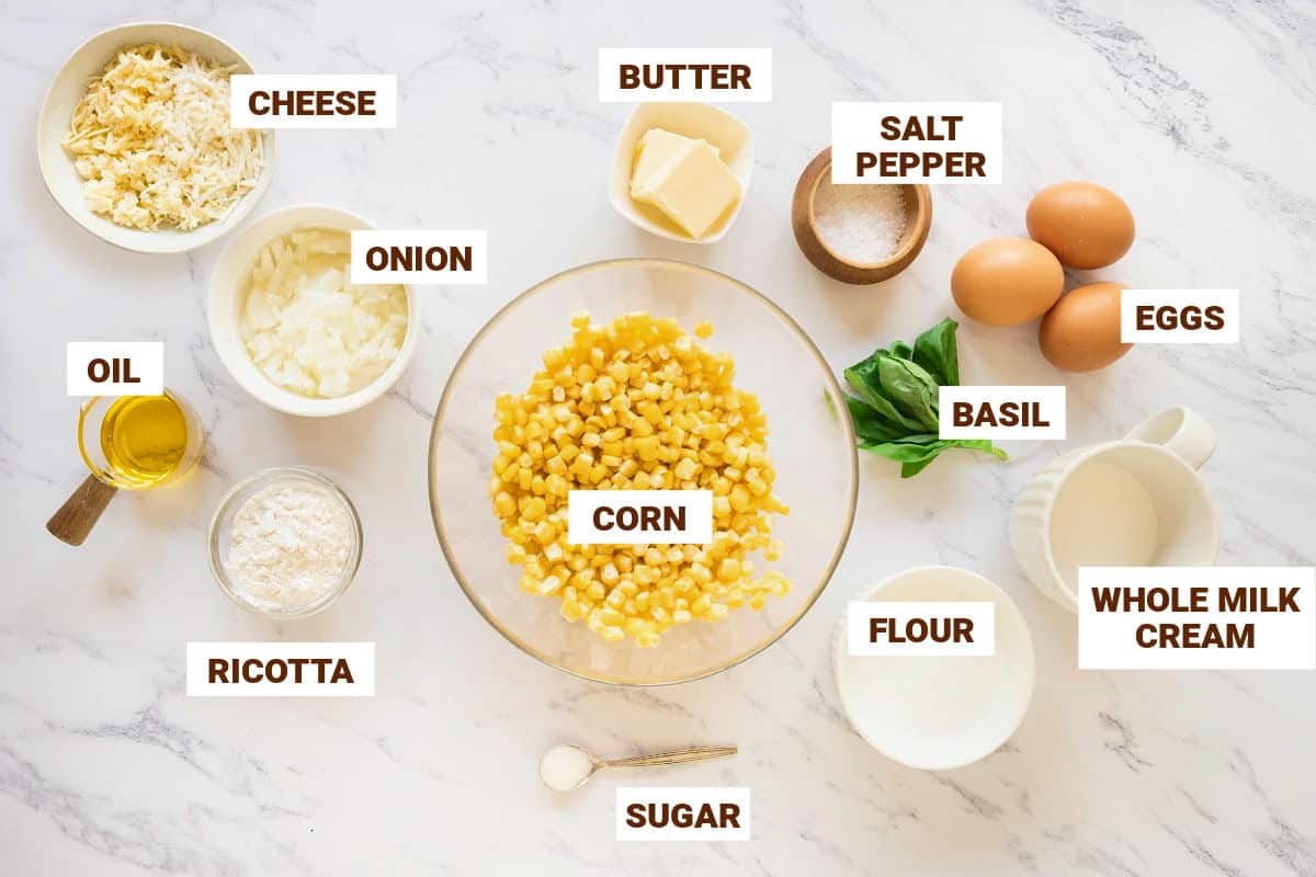 White marble with ingredients for corn pudding including cheese, basil, eggs, seasonings, cream and onion.