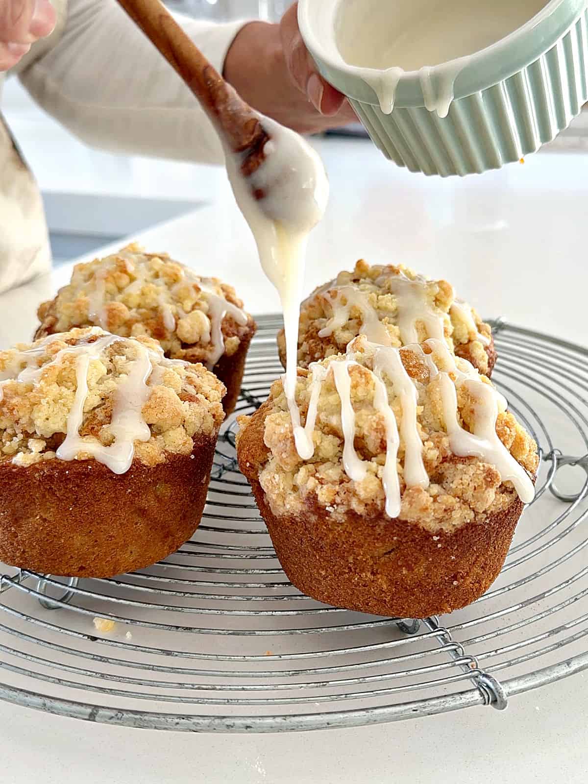 Glazing several apple crumble muffins on a wire rack, with a honey spoon.