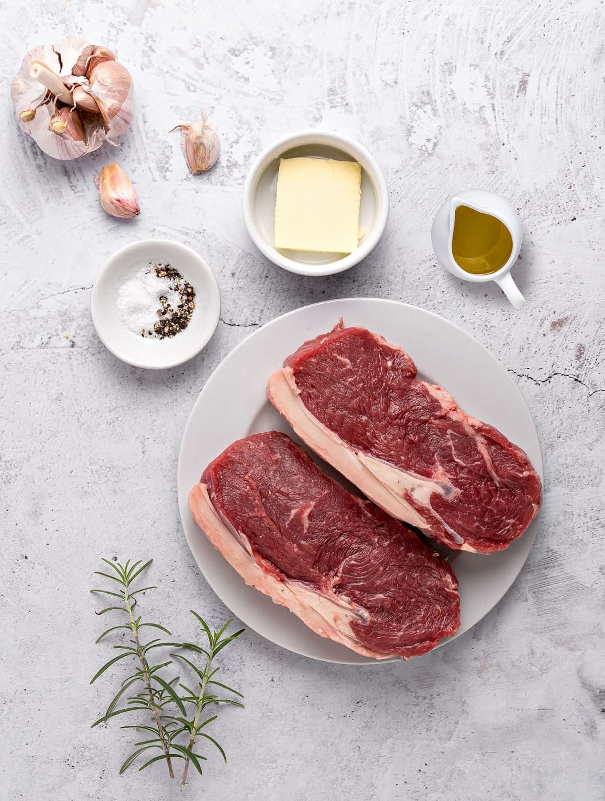 Ingredients for rosemary garlic steak on a light gray background, including butter, wine and seasonings.