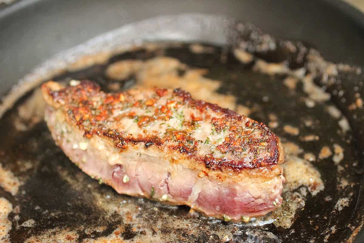 Half-cooked, seared steak on a dark pan with butter, rosemary and garlic.