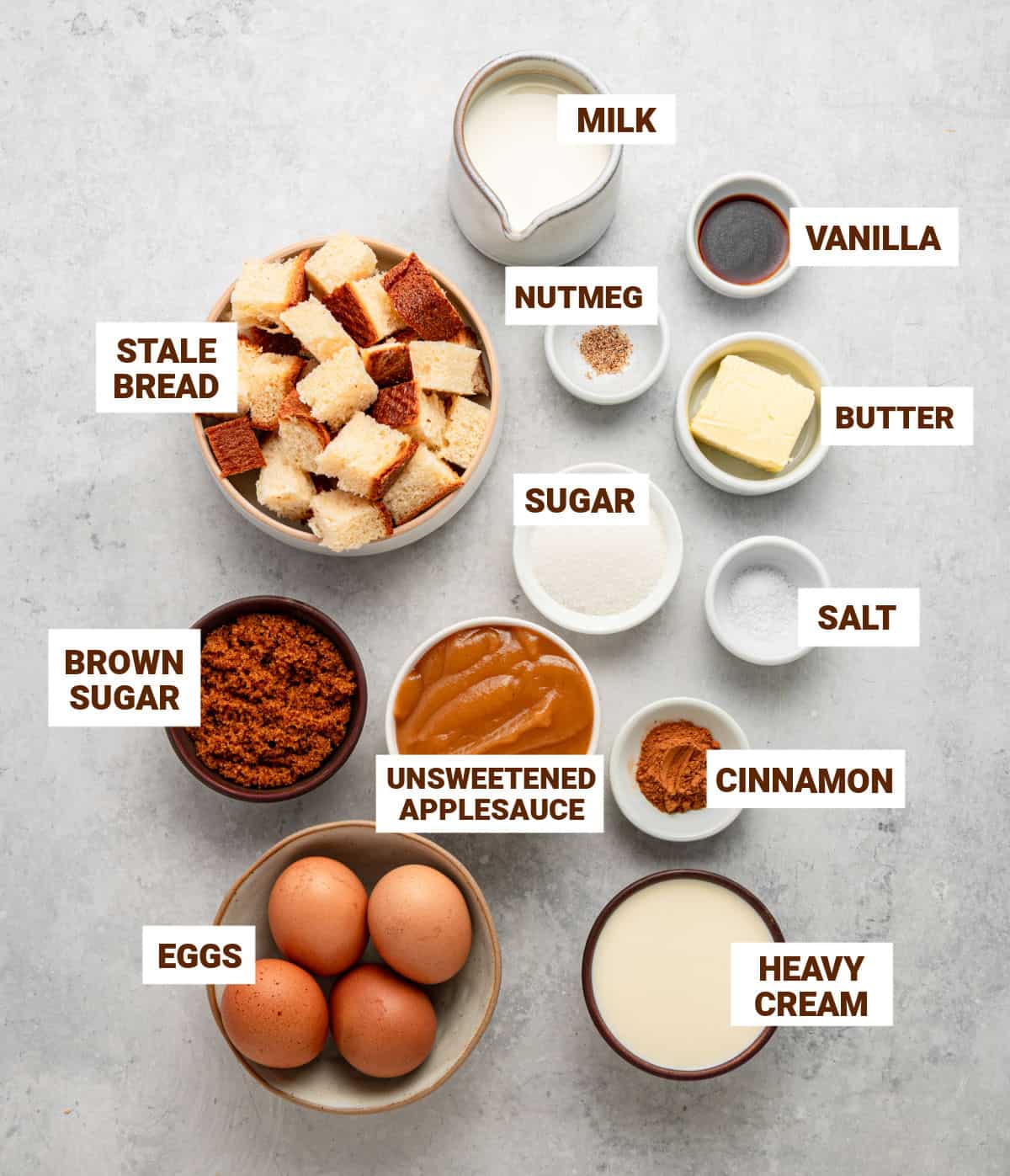 Ingredients for applesauce bread pudding in bowls on a gray surface, including cream, spices, butter, vanilla, brown sugar, eggs and milk.
