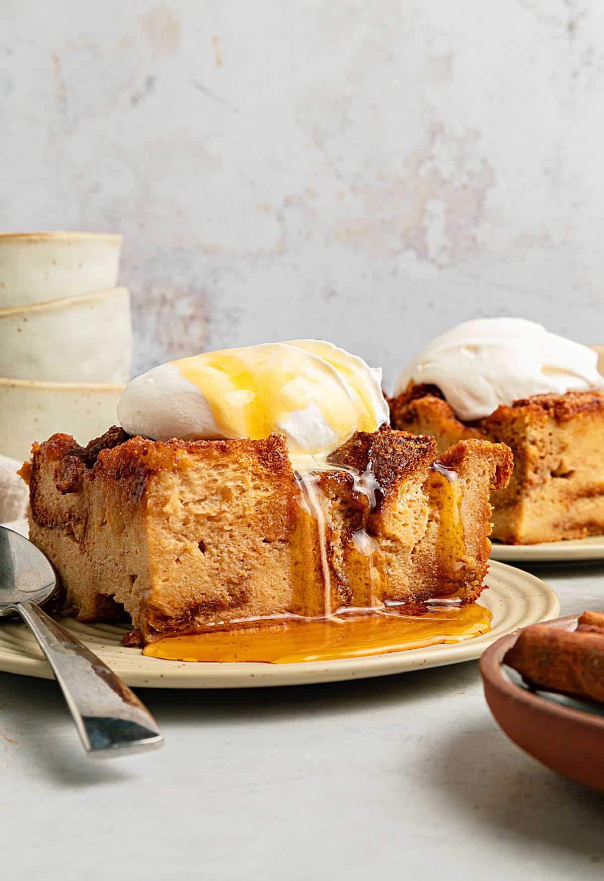 Light gray background with cream-topped bread pudding on beige plates. Maple syrup drizzle.
