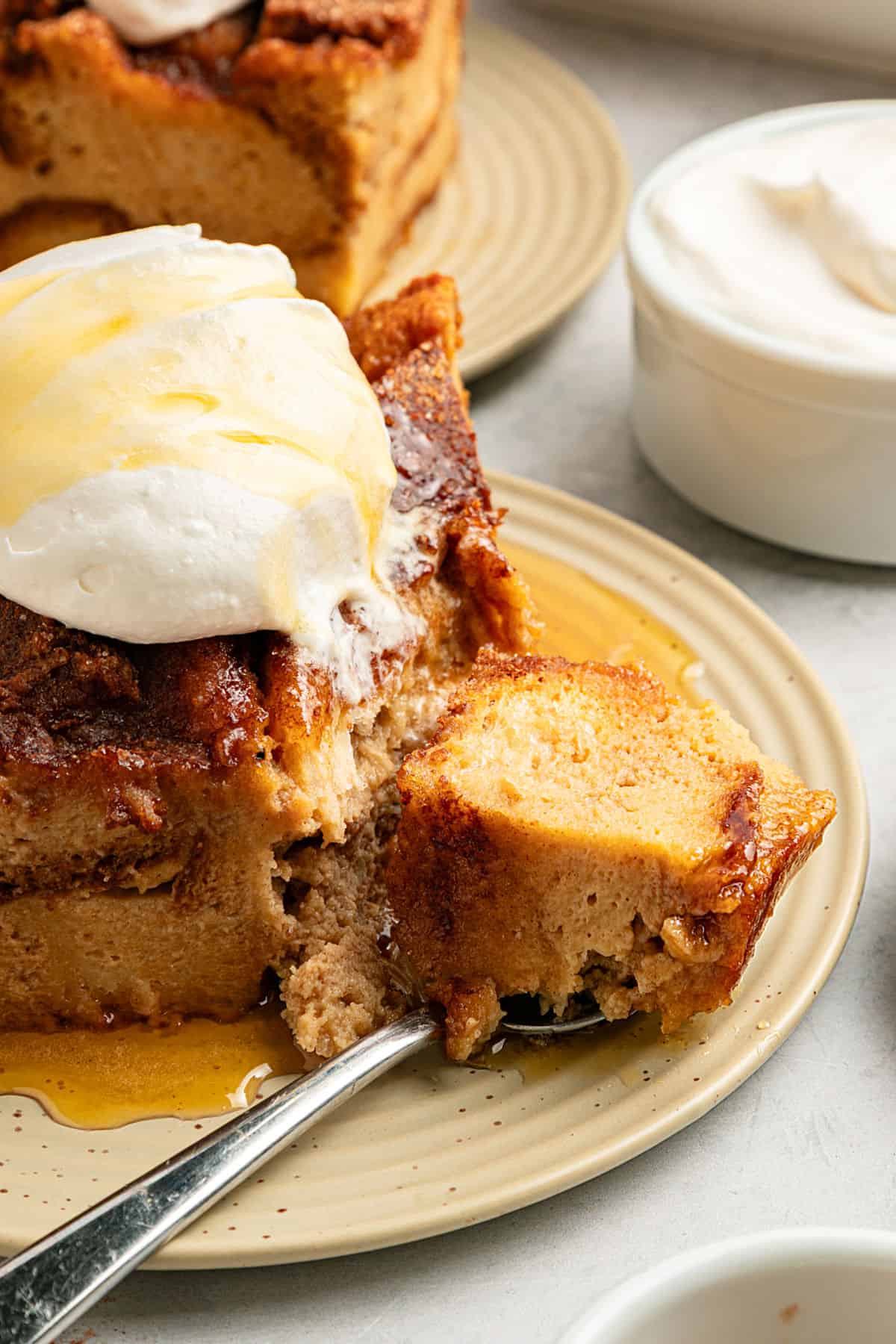 Partial view of beige plate with maple cream bread pudding slice. Silver spoon. Whipped cream bowl.