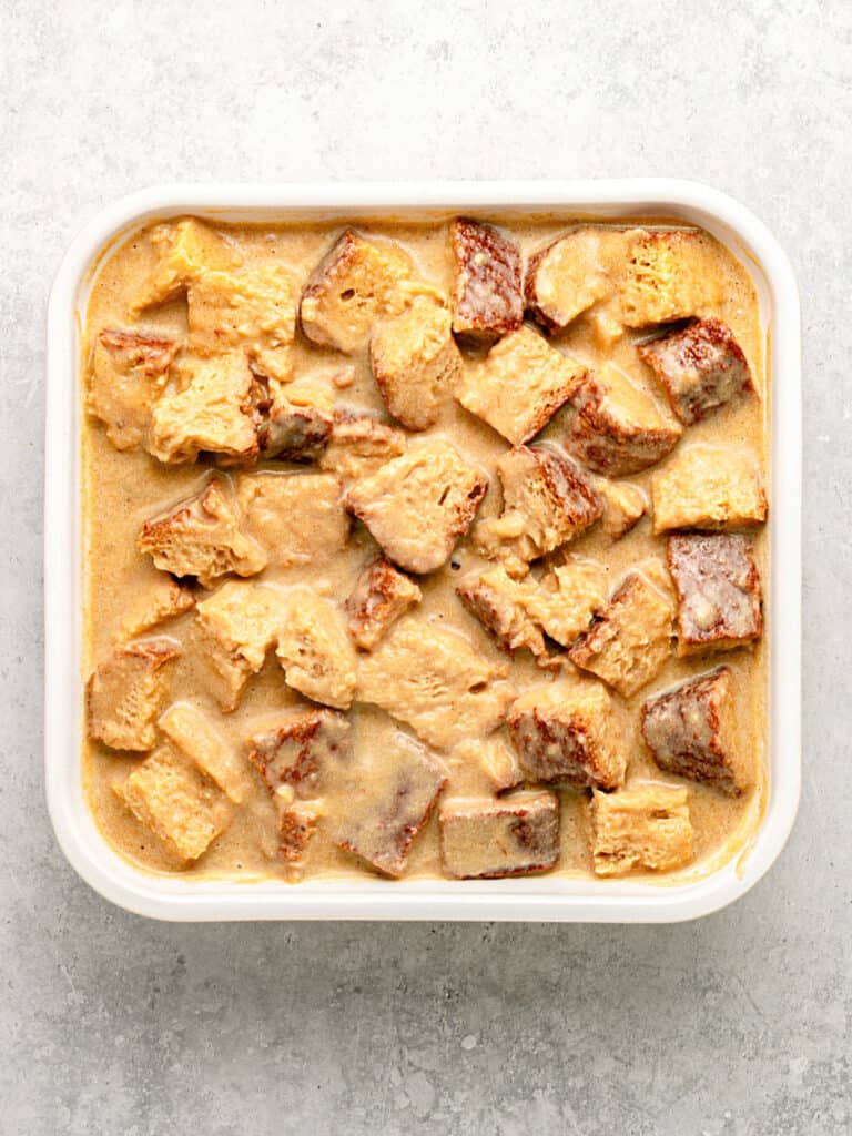 Square white baking dish with custard soaked brioche cubes. Light gray surface, top view.