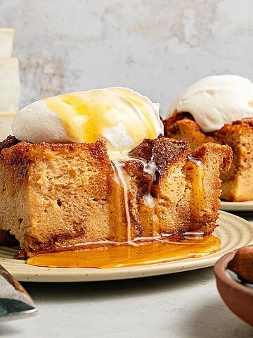 Maple and whipped cream-topped bread pudding servings on beige plates. Light gray background.