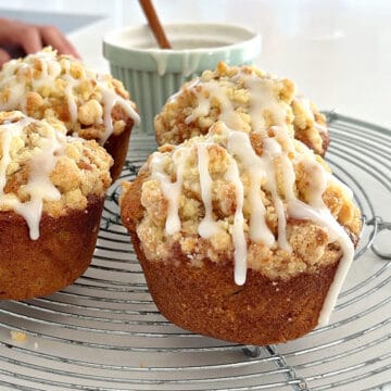 Wire rack with four glazed apple muffins with crumble. White surface, bowl with glaze.