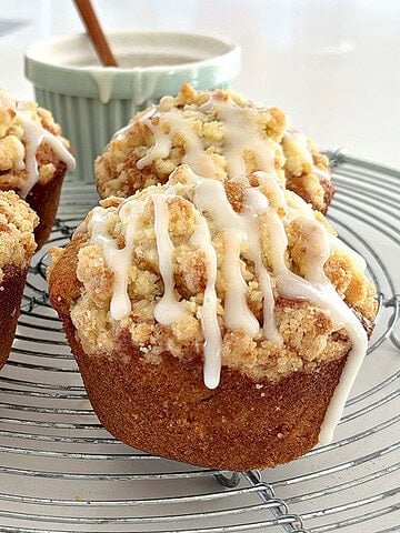 Wire rack with four glazed apple muffins with crumble. White surface, bowl with glaze.