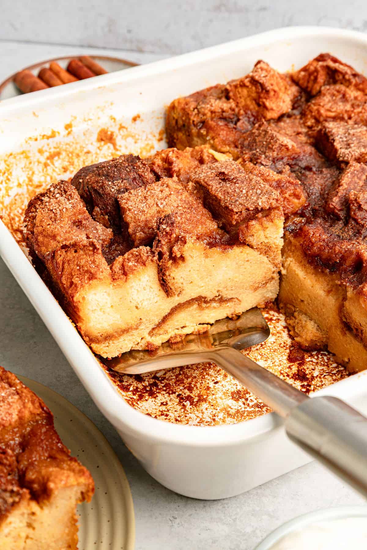 Metal spatula lifting slice of cinnamon-topped bread pudding. White baking dish, gray background.