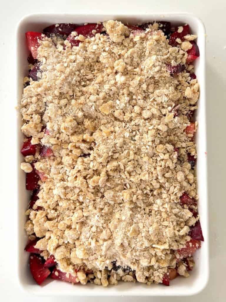 Oat crumble topped plum filling in a white rectangular baking dish. Top view.