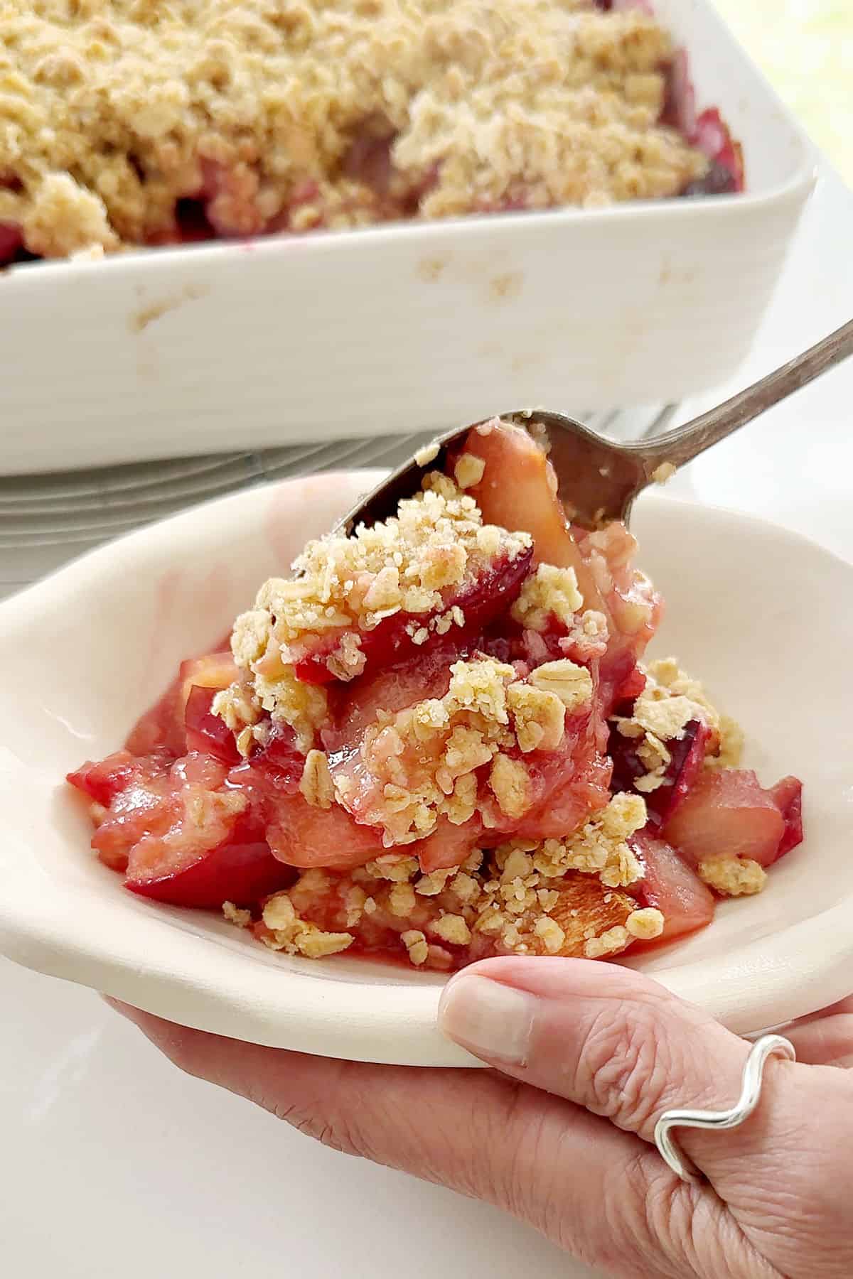 Spooning plum crisp on an oval cream-colored bowl. Baking dish in the background.