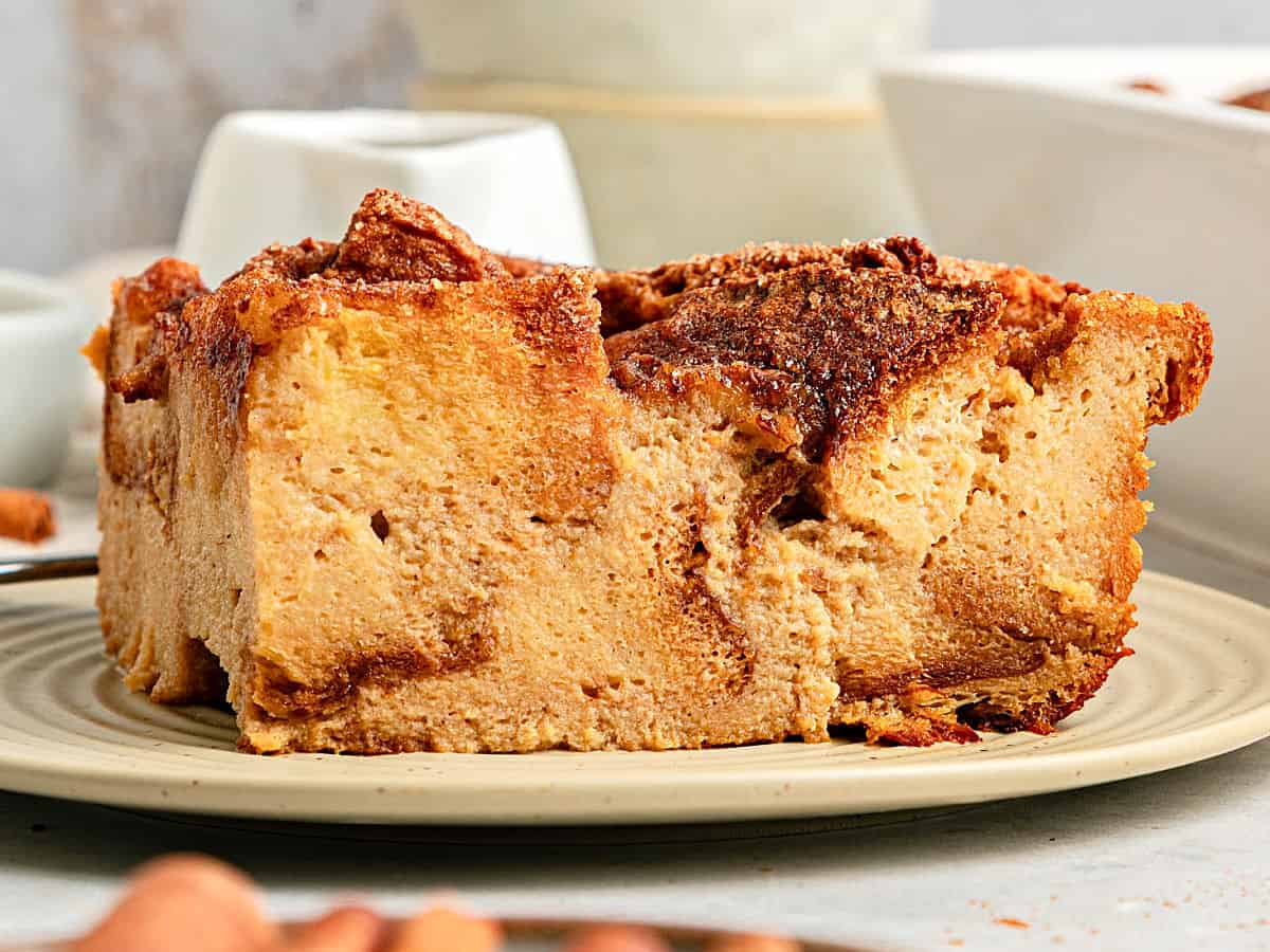 Close up serving of plain, creamy bread pudding on a beige plate. White bowls, gray background.