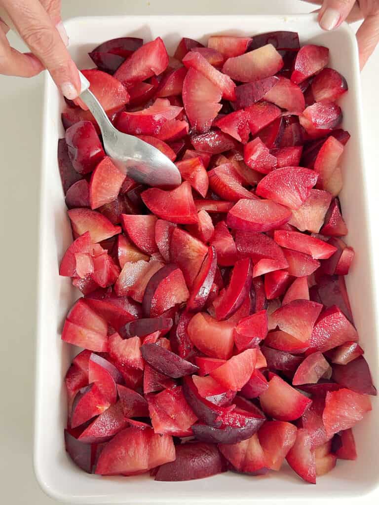 White rectangular baking dish with plum pieces. Hand with silver spoon.