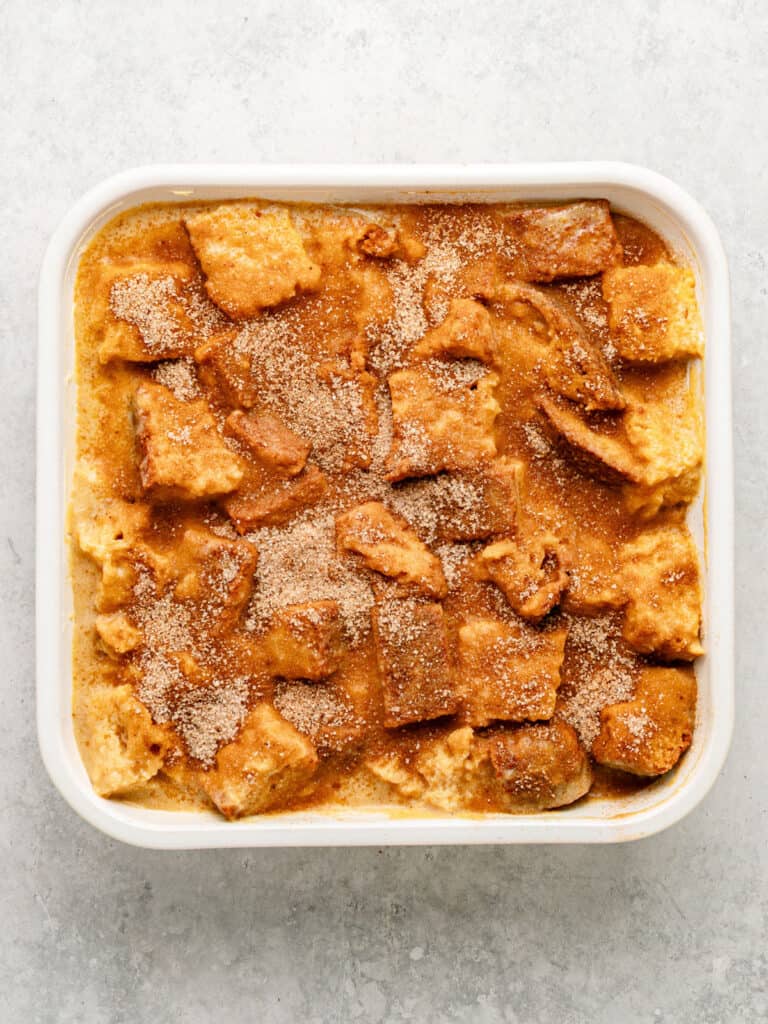 White square baking dish with cinnamon-topped custard soaked brioche cubes. Light gray surface, top view.