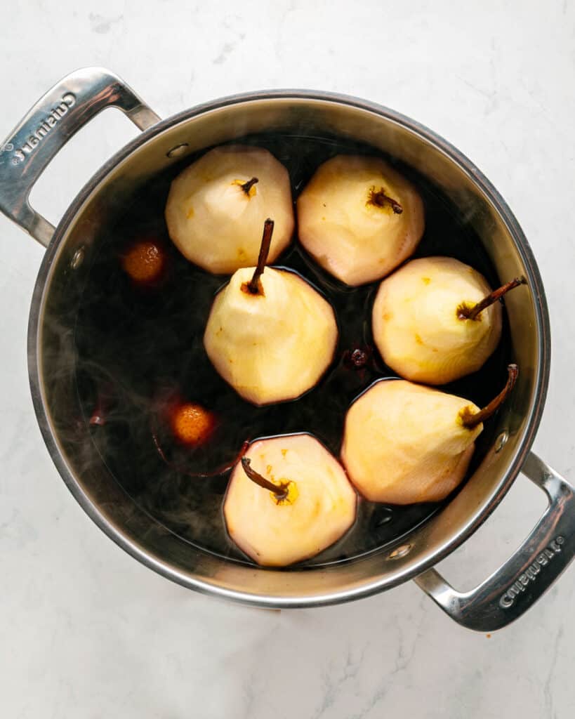 Metal saucepan with peeled whole pears and spiced red wine. White marbled surface.