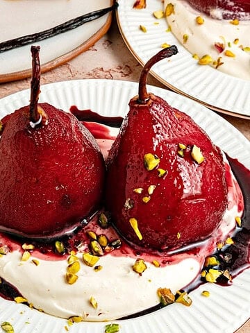 Whole spiced red wine poached pears on layer of whipped cream and syrup. White plate, pistachios.