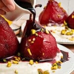 Dark red wine syrup poured over wine infused pears. White plate, whipped cream, chopped pistachios.