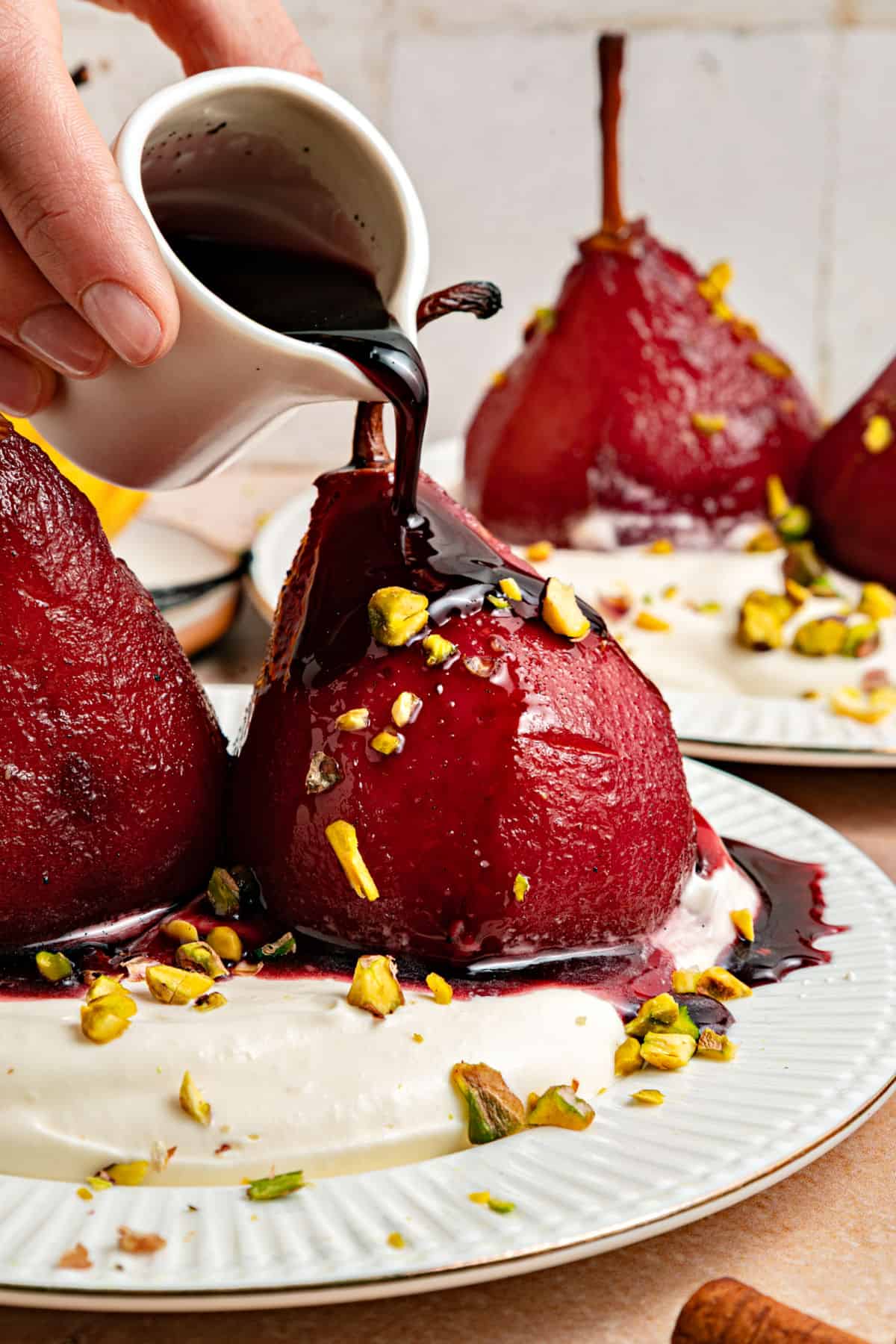 Spiced Red Wine Poached Pears with reduced wine sauce being poured. White plates, chopped pistachios.