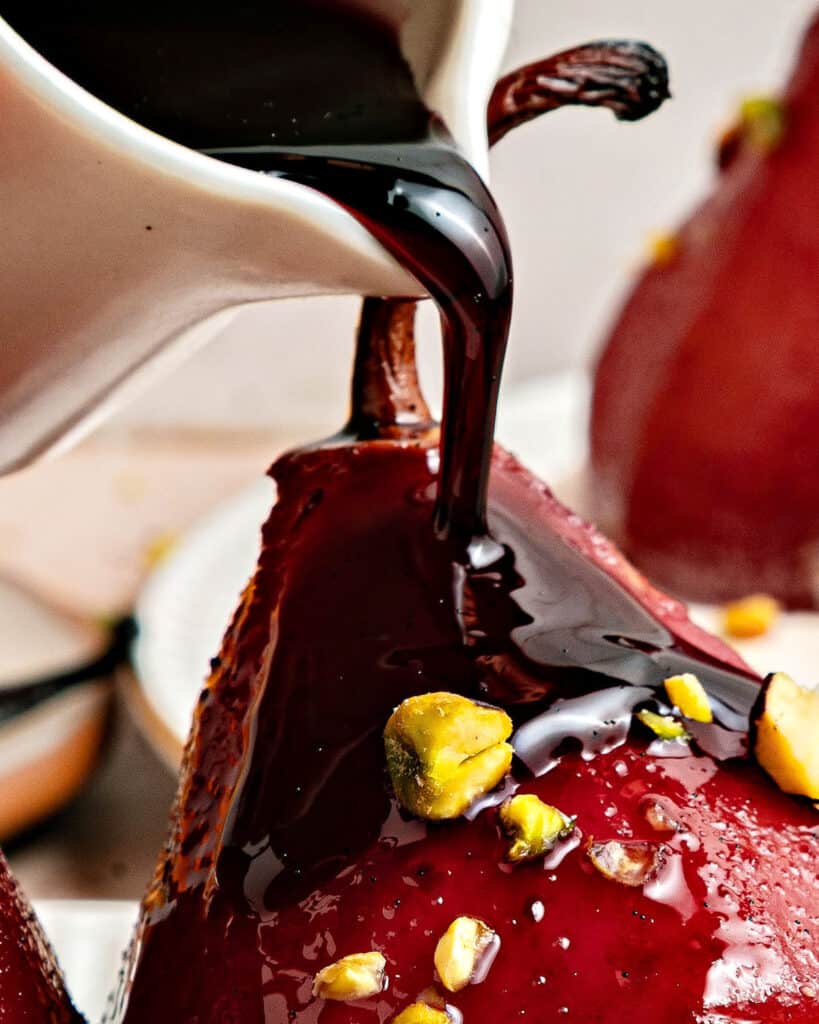 Close up pouring thick red wine sauce from a white jar onto poached pears.