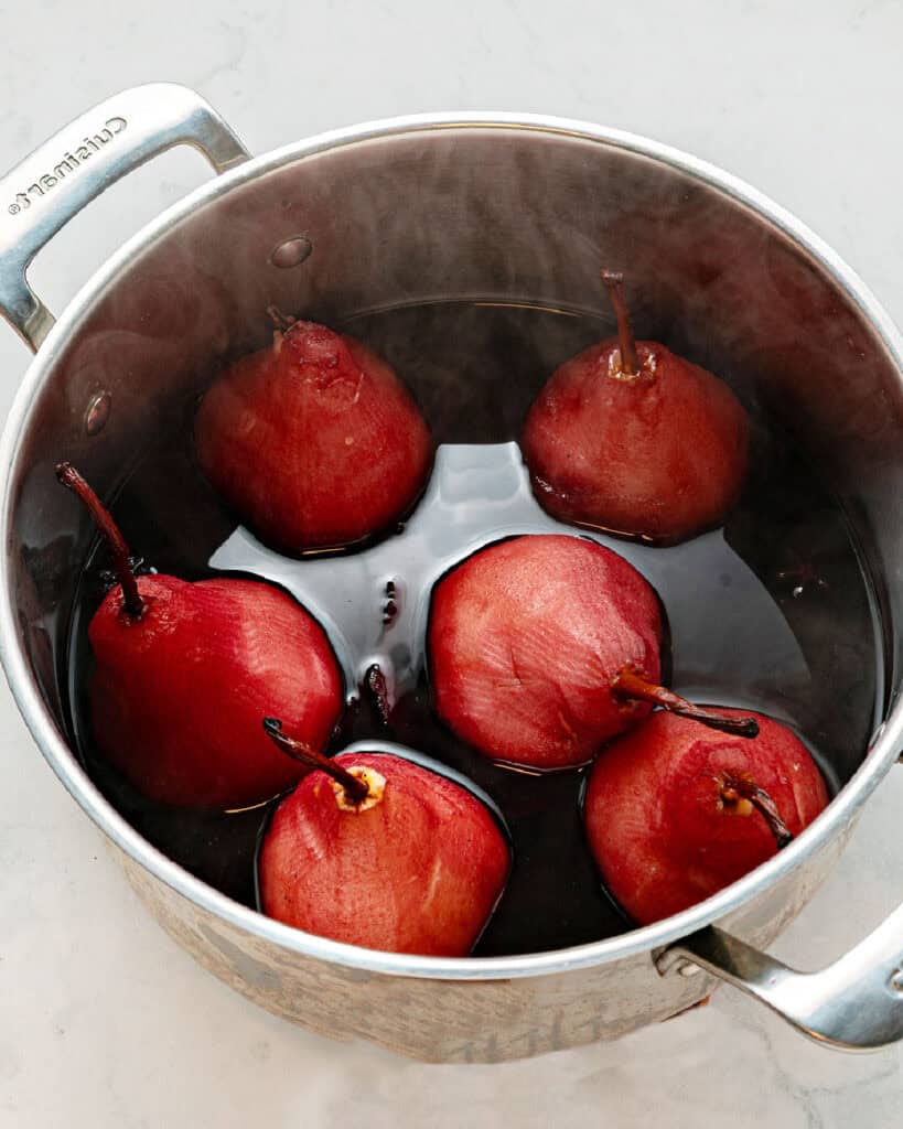 Six whole red wine poached pears in a pot with spiced wine liquid.