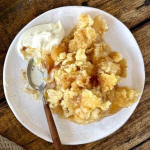 Vintage apple dump cake serving on a white plate with cream. A spoon. Wooden surface.