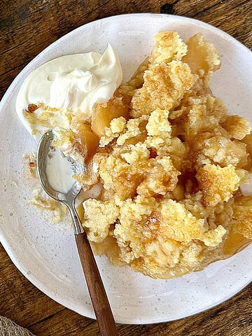 Vintage apple dump cake serving on a white plate with cream. A spoon. Wooden surface.
