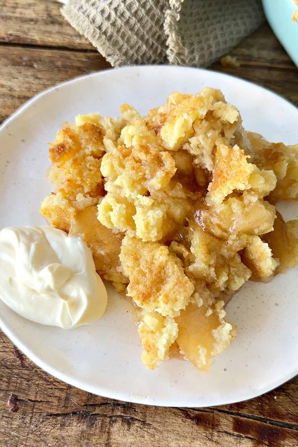 White plate with apple dump cake serving and cream. Wooden surface.